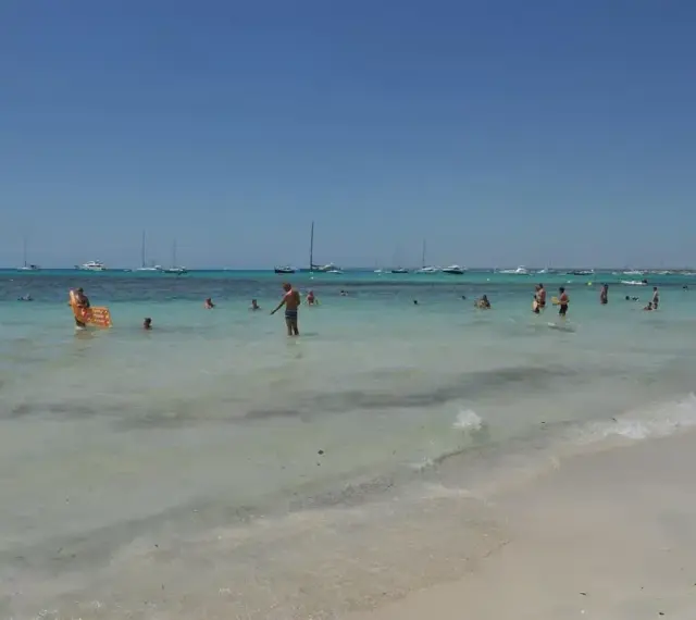 Słoneczny dzień na plaży Es Trenc. Ludzie kąpią się w turkusowej wodzie, a na horyzoncie widać żaglówki.