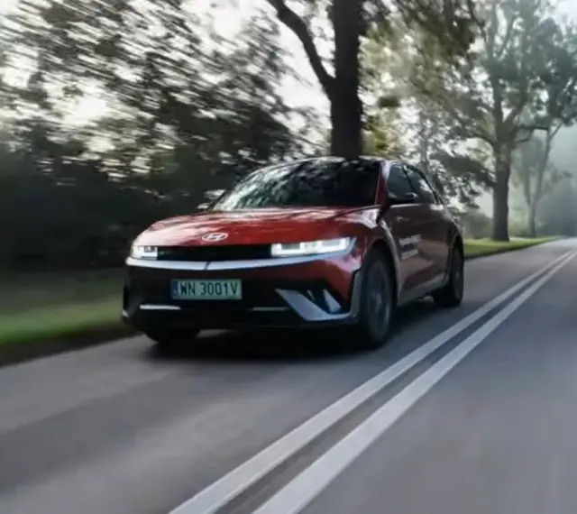 Czerwony Hyundai Ioniq 5 jedzie po wiejskiej drodze wśród drzew.