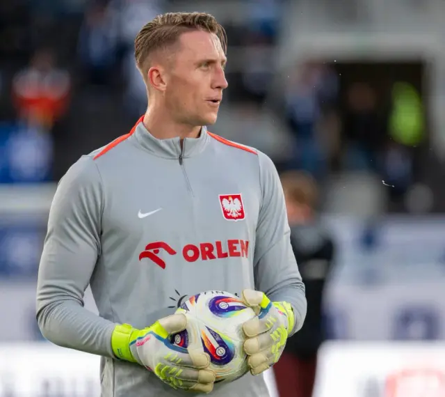 Bramkarz w szarej bluzie z orłem i napisem ORLEN, trzymający piłkę. Kolejny z polscy piłkarze za granicą, gotowy do gry.
