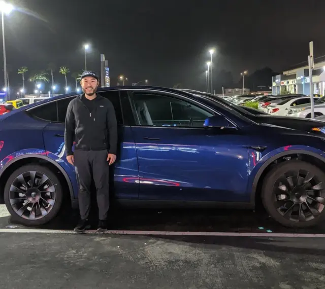 Mężczyzna stoi obok ciemnoniebieskiego Tesli Model Y na nocnym parkingu.