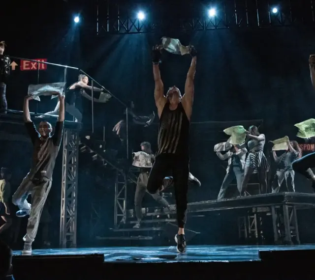 Na scenie "Metro Janusza Józefowicza" tancerze wznoszą gazety, tworząc dynamiczną choreografię.