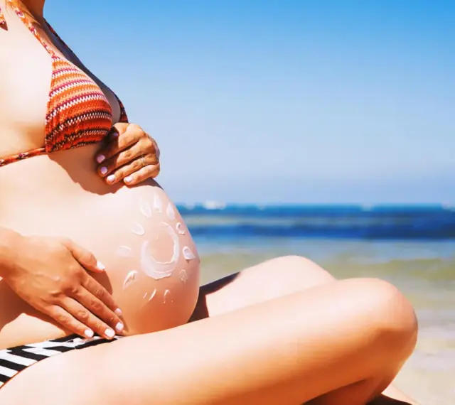 Ciężarna kobieta na plaży, z kremem przeciwsłonecznym na brzuchu w kształcie słońca. Bezpieczne opalanie natryskowe w ciąży.