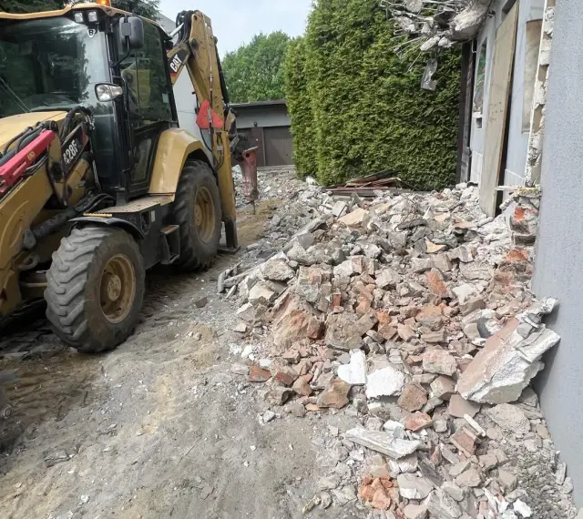 Żółta koparko-ładowarka pracuje przy wyburzeniu domu. Gruz i cegły leżą na ziemi. Koszt wyburzenia domu jest wysoki.