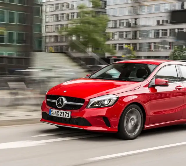 Czerwony Mercedes-Benz W176 pędzi ulicą miasta, mijając nowoczesne budynki.