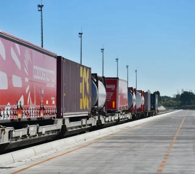 Długi pociąg towarowy z kontenerami, w tym czerwonymi z logo COSCO i żółtymi, czeka na dalszy transport kolejowy.
