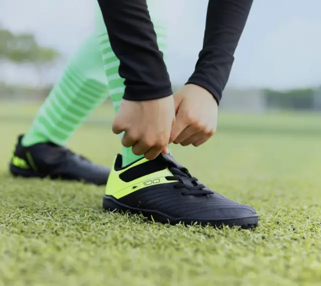 Dłonie wiążą sznurówki czarnych butów sportowych z neonowożółtymi wstawkami na zielonej murawie. To pierwszy krok, by dowiedzieć się, jak rozciągnąć buty sportowe.