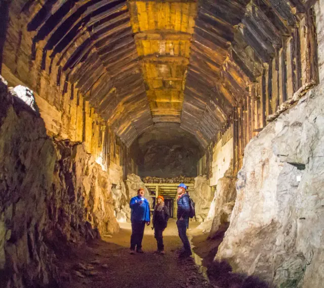 Zwiedzanie podziemnego kompleksu Projekt Riese. Trzech turystów w kaskach oświetla latarkami mroczny tunel z drewnianymi stropami.