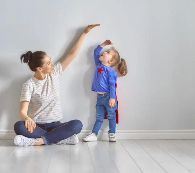 Mama mierzy wzrost córeczki w stroju superbohaterki. To właśnie wtedy dzieci rosną najszybciej, zaskakując nas każdego dnia.