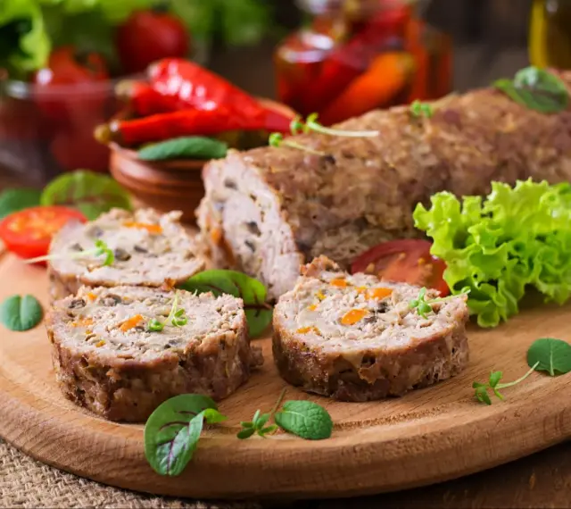 Pokrojone zrazy z mięsa mielonego z warzywami na desce, obok sałata, pomidory i papryczki chili.