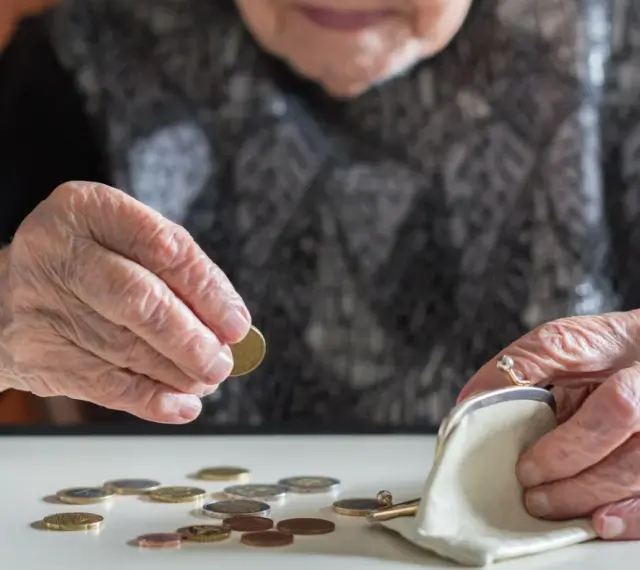 Dłoń starszej kobiety z pierścionkiem wkłada monetę do portmonetki. Rozrzucone monety na stole sugerują trudności finansowe, być może związane z emeryturą po mężu.