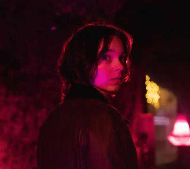 Młoda kobieta z zaciekawieniem spogląda przez ramię w mrocznym, neonowym otoczeniu. Czy to scena z nowego serialu Netflix?