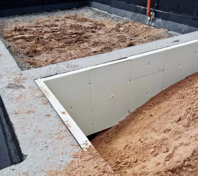 Fundament pod altanę w budowie: betonowe ławy, izolacja ze styropianu i nasypy ziemi. Słupki fundamentowe pod altanę czekają na montaż.