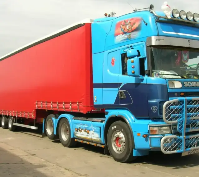 Naczepa jumbo w jaskrawym kolorze czerwonym, ciągnięta przez niebieską Scanię z malowidłami.