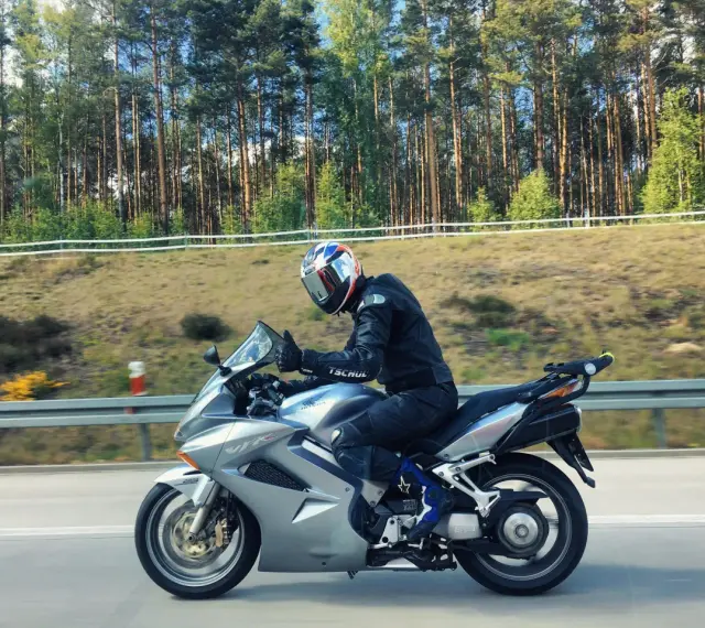 Motocyklista w kasku jedzie na srebrnym Honda VFR 800 wzdłuż leśnej drogi.