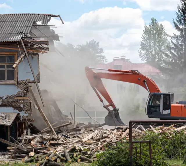 Koparka burzy zniszczony budynek, unosząc tumany kurzu. W tle inne domy i drzewa. Rozbiórka budynku formalności w toku.