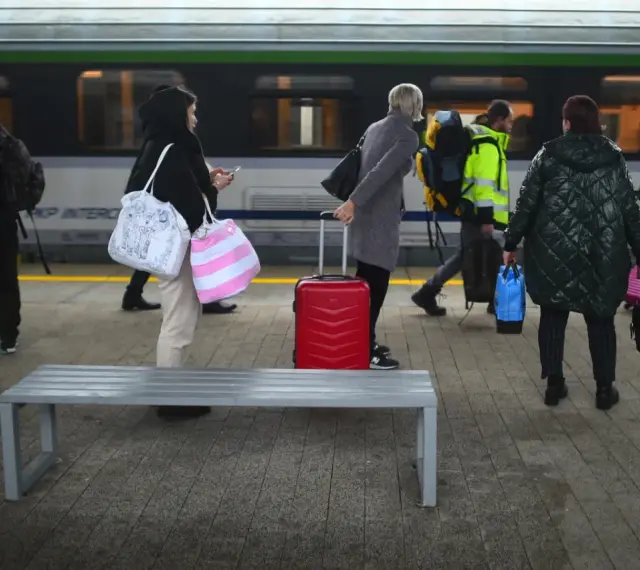 Ludzie z bagażami czekają na peronie PKP Intercity. Czerwona walizka stoi obok ławki.