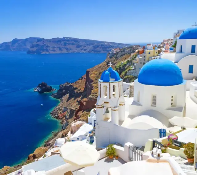 Santorini wycieczki: białe domy z niebieskimi kopułami na klifie nad błękitnym morzem. Statek płynie w oddali.