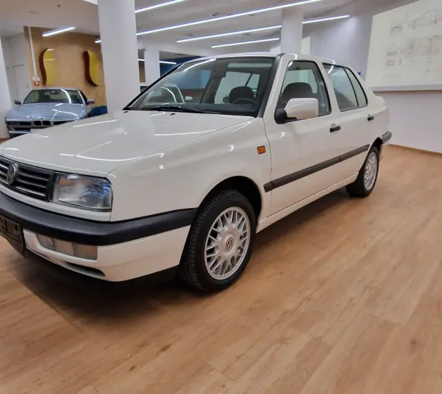 Biały Volkswagen Vento z lat 90. stoi na drewnianej podłodze w salonie samochodowym.