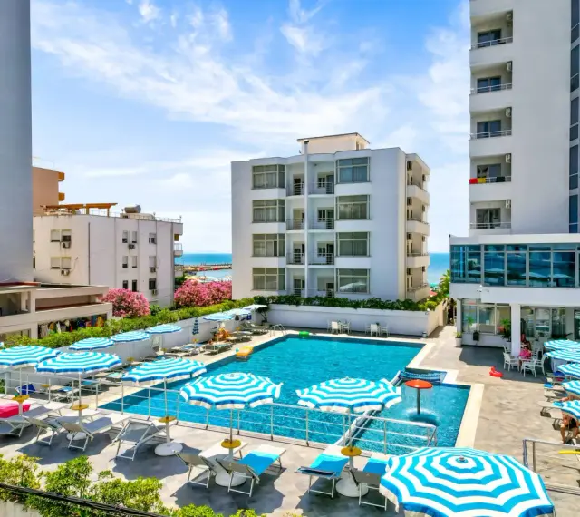 Albania - wczasy samolotem all inclusive. Basen hotelowy z leżakami i parasolami, w tle budynki i morze. Idealne miejsce na relaks.