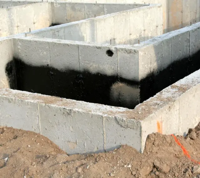 Surowe ściany fundamentowe z betonu, częściowo pokryte czarną izolacją przeciwwilgociową, otoczone ziemią.