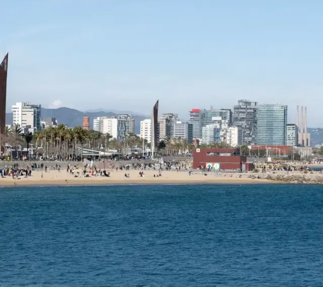 Ludzie odpoczywają na plaży, a w tle widać nowoczesne budynki Barcelony. To miasto leży nad Morzem Śródziemnym.