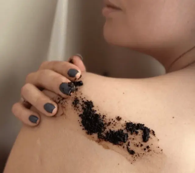 Dłoń z ciemnym lakierem nakłada peeling kawowy na ramię. Efekty po miesiącu regularnego stosowania są widoczne.