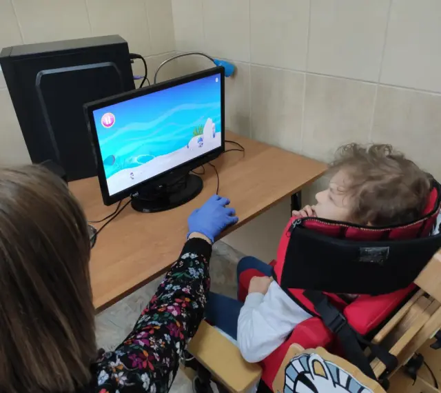 Tyflopedagog w niebieskiej rękawiczce pomaga dziecku w specjalnym foteliku grać w grę na komputerze.