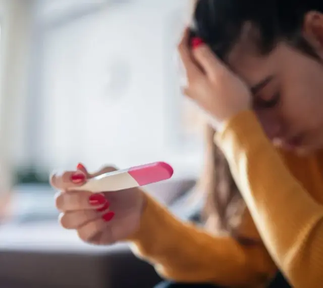 Kobieta z opuszczoną głową trzyma test ciążowy. Jej dłoń z czerwonym lakierem do paznokci zaciska się na teście. Tokofobia może wywoływać takie emocje.
