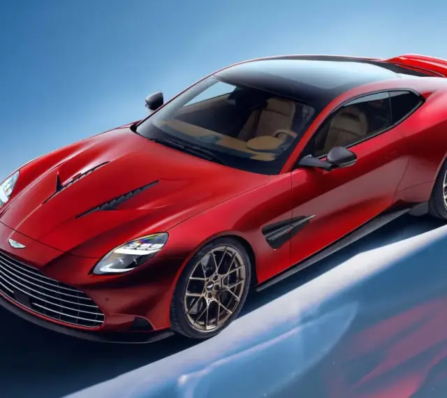 Czerwony Aston Martin Vanquish z brązowym wnętrzem i czarnym dachem, prezentujący swoje sportowe linie na tle błękitnego nieba.