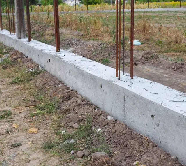 Zbrojony fundament czeka na wylewanie betonu. Widać stalowe pręty wystające z betonowej ławy fundamentowej.