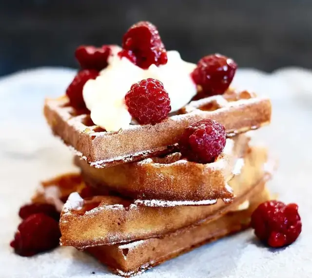 En stapel gyllene belgiska våfflor toppade med vispgrädde och färska hallon, pudrade med florsocker. En härlig frukost eller efterrätt.