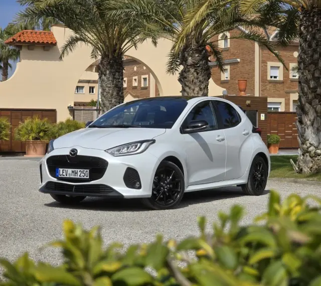 Biała Mazda 2 Hybrid zaparkowana przed budynkiem z palmami. Stylowy miejski samochód gotowy na podróż.