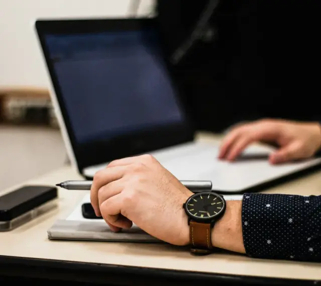Dłoń z zegarkiem trzyma długopis nad dokumentem, obok laptop. Czas na skargę na przewlekłość postępowania opłatę.