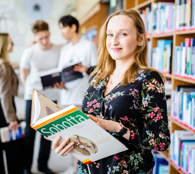 Studentka na Uniwersytecie Wrocławskim, kierunek prawo, przegląda atlas anatomii. W tle inni studenci i regały z książkami.