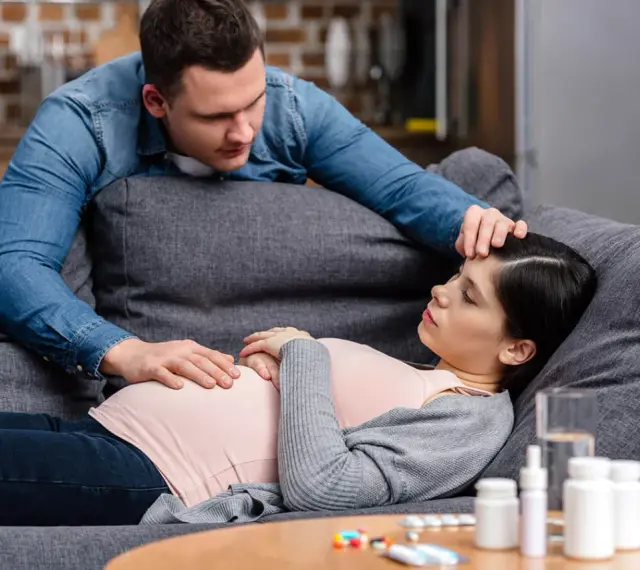 Zmartwiony partner troskliwie dotyka czoła ciężarnej kobiety, która leży na kanapie. Na stoliku leki, sugerujące problemy zdrowotne, być może angina w ciąży.