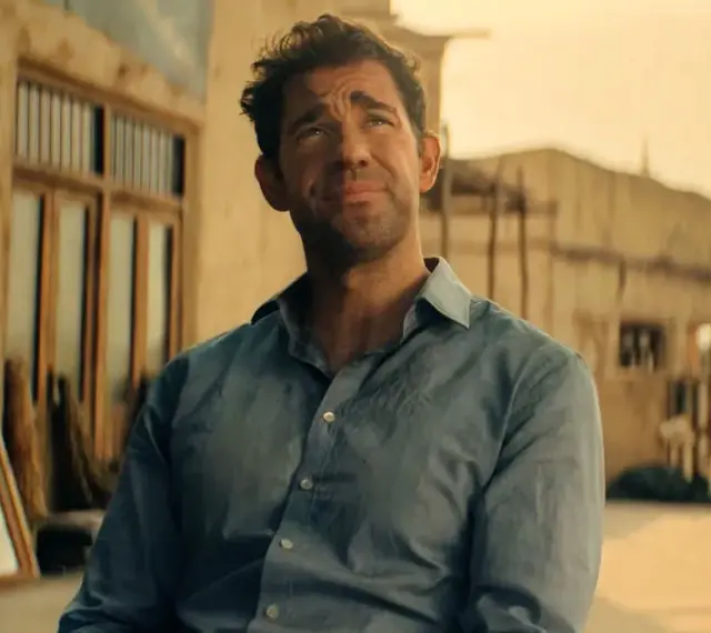 John Krasinski w niebieskiej koszuli, zamyślony. Widzimy fragmenty jego filmów, seriali i programów.