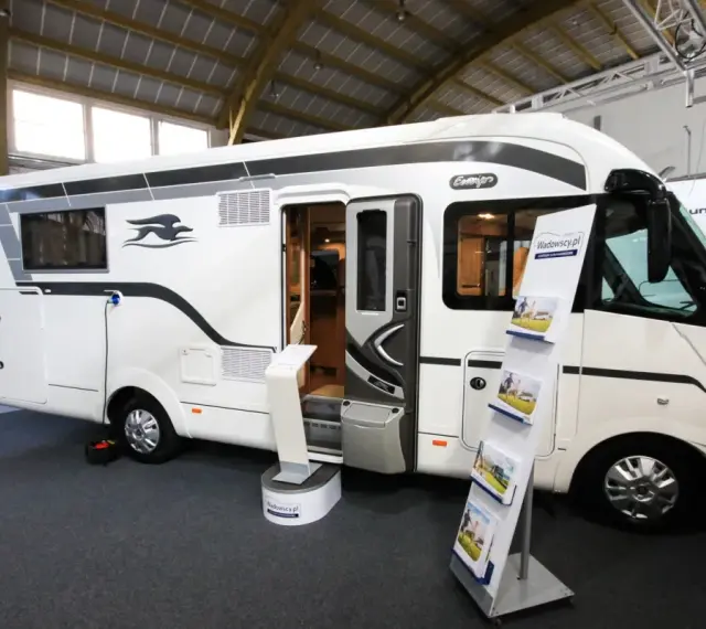 Biała kamperka z otwartymi drzwiami na targach caravaningowych Poznań. W środku widać wyposażenie, a na zewnątrz stojaki z ulotkami.