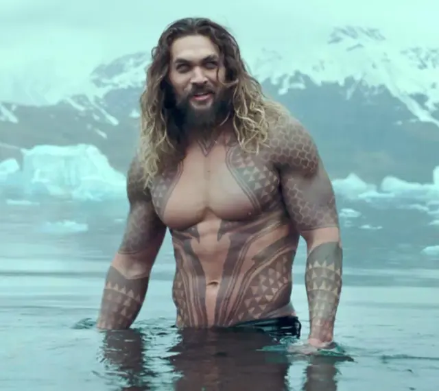 Jason Momoa w wodzie, z tatuażami, jak w filmach i serialach. W tle góry i lód.