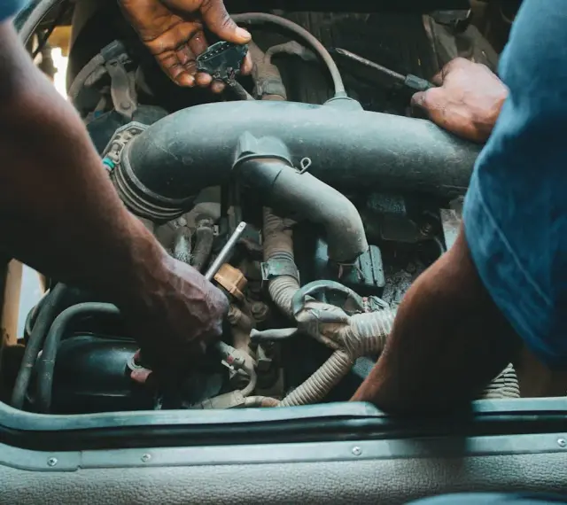 Mechanicy pracują przy silniku, jeden z nich trzyma wtyczkę, drugi dokręca śrubę. Może to być naprawa zaworu par paliwa.