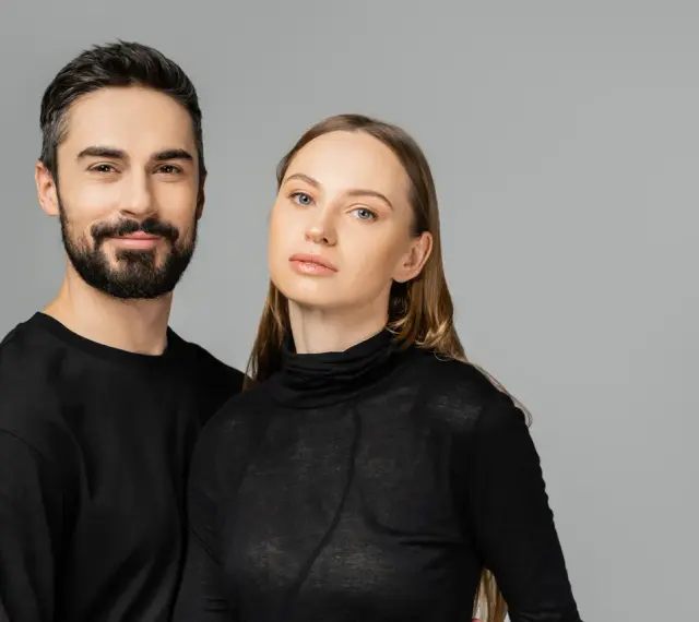 Para, która wygląda jakby mogła nosić ubrania unisex. Mężczyzna z brodą i kobieta z długimi włosami, oboje w czarnych, prostych ubraniach.