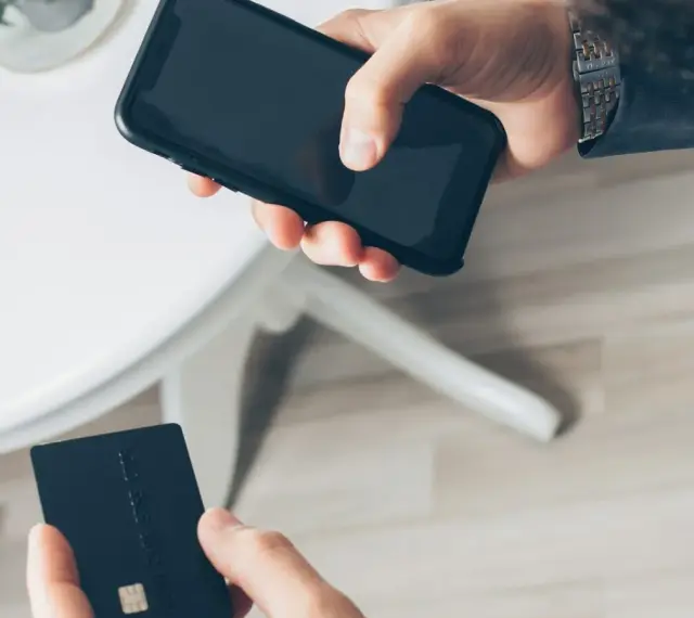 Dłoń trzyma smartfon, druga dłoń czarną kartę płatniczą. Szybkie płatności zbliżeniowe, jak w PKO BP, to wygoda.