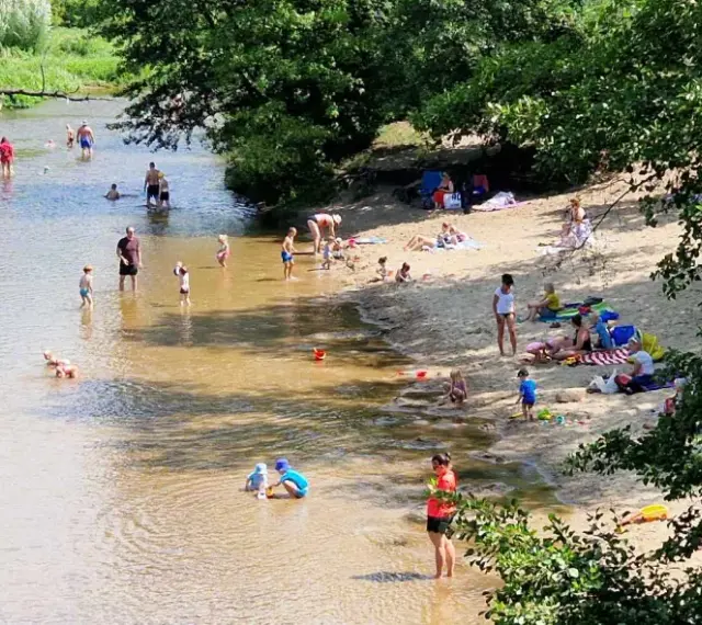 Słoneczny dzień nad Świdrem. Ludzie odpoczywają na piaszczystej plaży i kąpią się w rzece. Dzieci bawią się w wodzie.