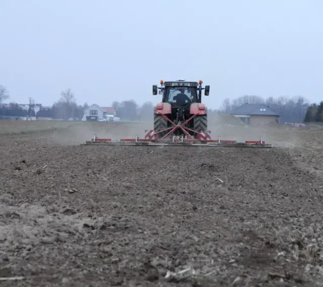 Czerwony traktor z agregatem uprawowym przygotowuje pole pod zasiew. Dzierżawa ziemi w pełni.