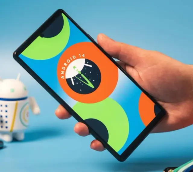 Dłoń trzyma smartfon z logo Android 14. Obok stoi figurka robota.