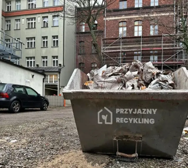 Kontener na śmieci z napisem "Przyjazny Recykling" wypełniony papierem. W tle budynki i czarny samochód. Data: 17 09 04.