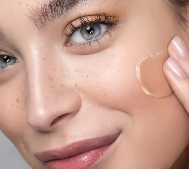 Delikatna mastyka na policzku kobiety, która rozprowadza ją palcem. Skóra z piegami i subtelnym makijażem.