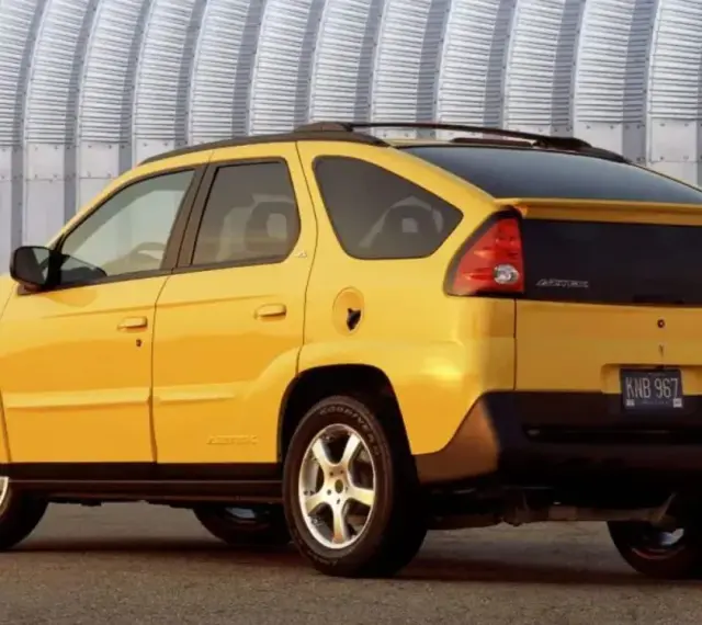 Żółty Pontiac Aztek z charakterystycznym tyłem i spojlerem, zaparkowany przed falistą, metalową ścianą.