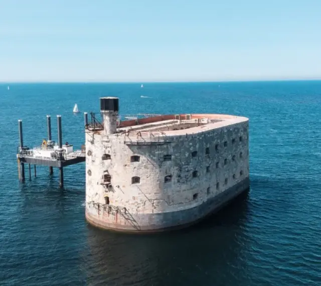 Fort Boyard, potężna twierdza na morzu, otoczona błękitną wodą. Obok platforma wiertnicza i żaglówka.
