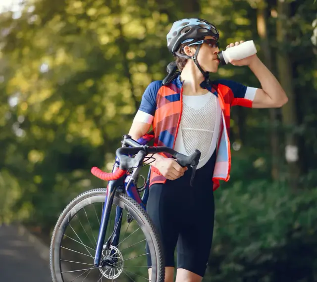 Kolarz w kasku i stroju sportowym pije wodę, trzymając rower. Dba o nawodnienie i dostarczenie makroskładników po wysiłku.