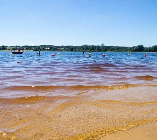 Słoneczny dzień nad zalewem. Ludzie kąpią się, pływają łódką, a czerwone i żółte boje wyznaczają bezpieczną strefę.